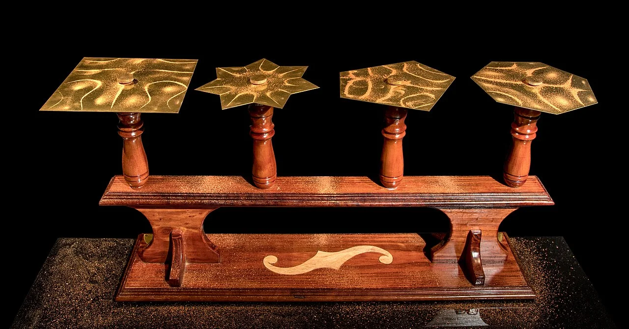 Four brass Chladni plates on wooden stands, their sand arranged into geometric vibration patterns